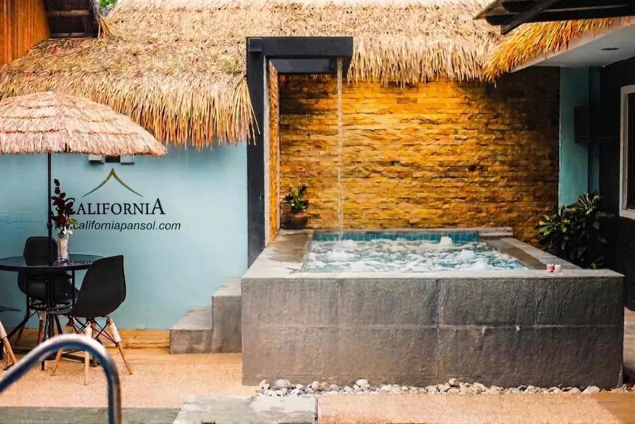 Villa 1 — raised square stone jacuzzi with a wall spout beside a thatch umbrella and the branded 'California / californiapansol.com' wall